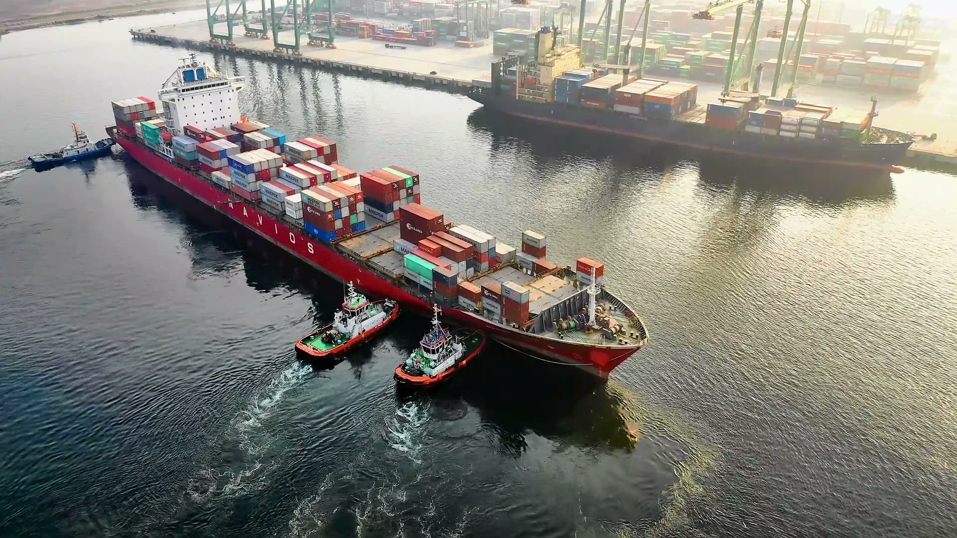 Aerial view of a fully-loaded container ship being escorted by tugboats into a UK port — gantry cranes lining the dockside